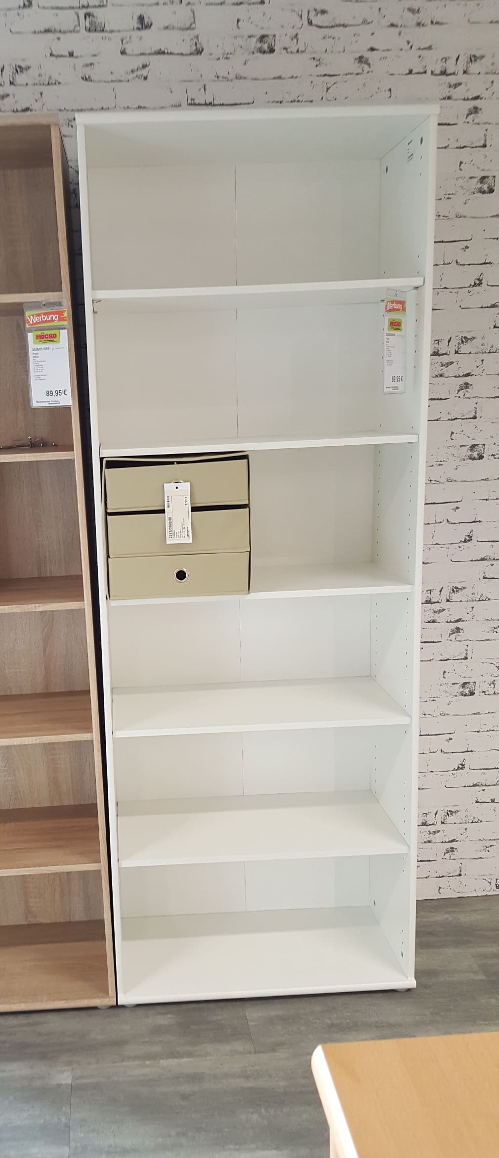 Das Regal Umut: ein weißes Bücherregal mit fünf Fächern, von denen eines zwei beigefarbene Kisten enthält, an einer weißen Backsteinwand.