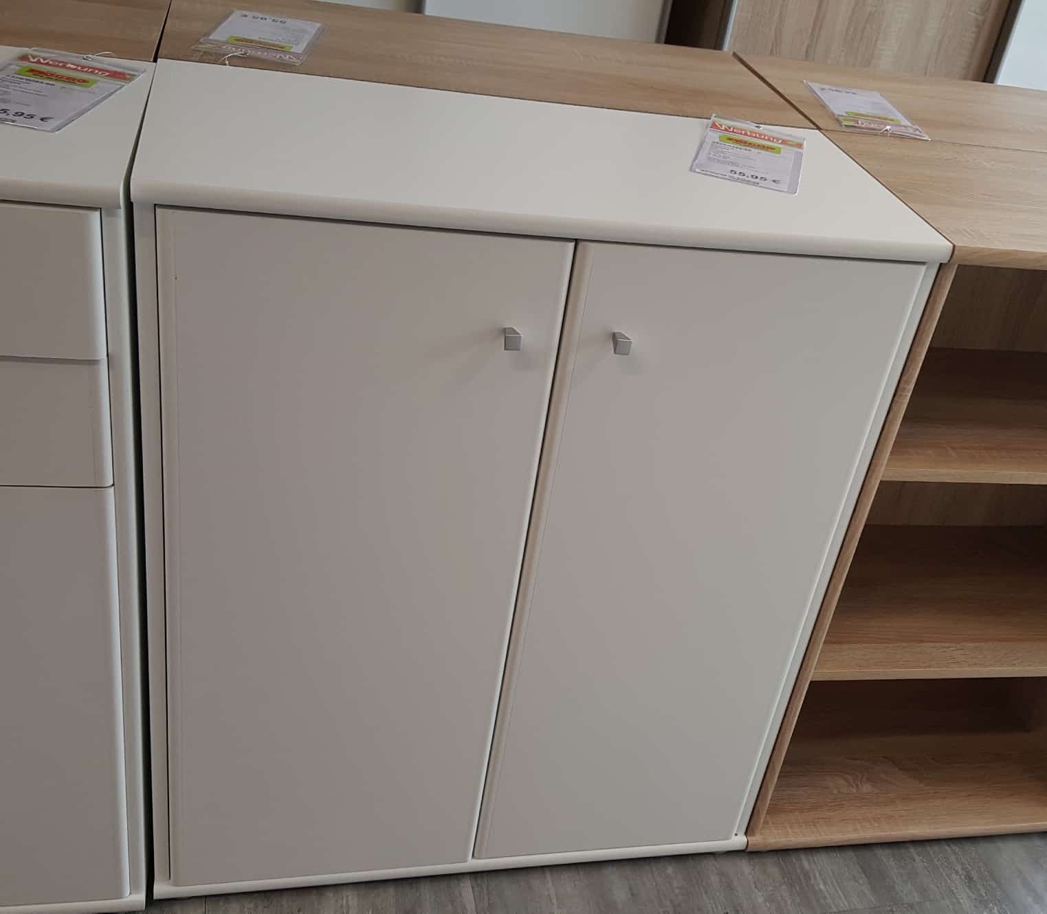 Die Kommode Umut ist ein weißer Schrank mit zwei Türen und silbernen Griffen, der mit anderen Holzmöbeln gezeigt wird.