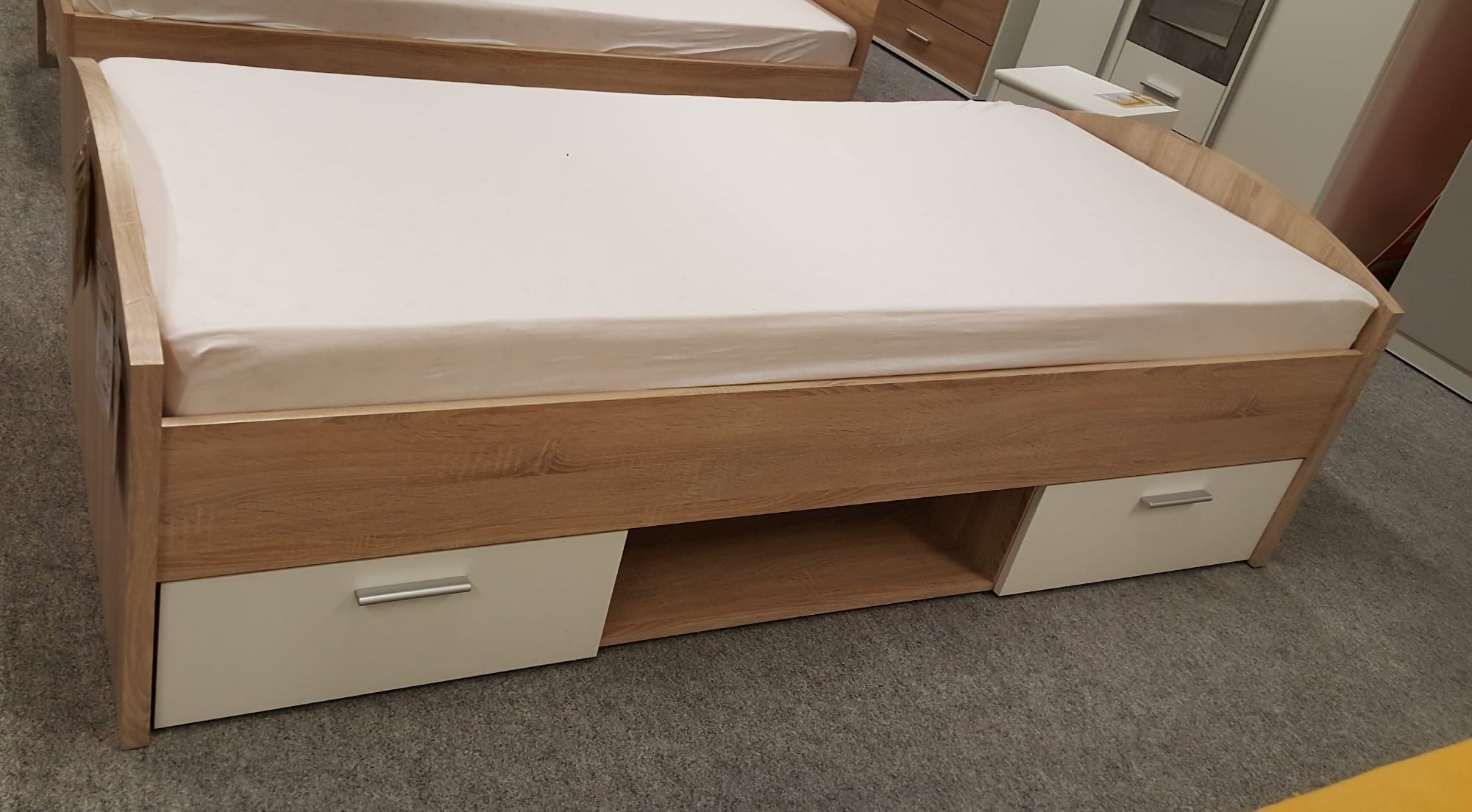 Jugendbett Yolande mit weißer Matratze und zwei Schubladen, auf einem grauen Teppich aufgestellt.