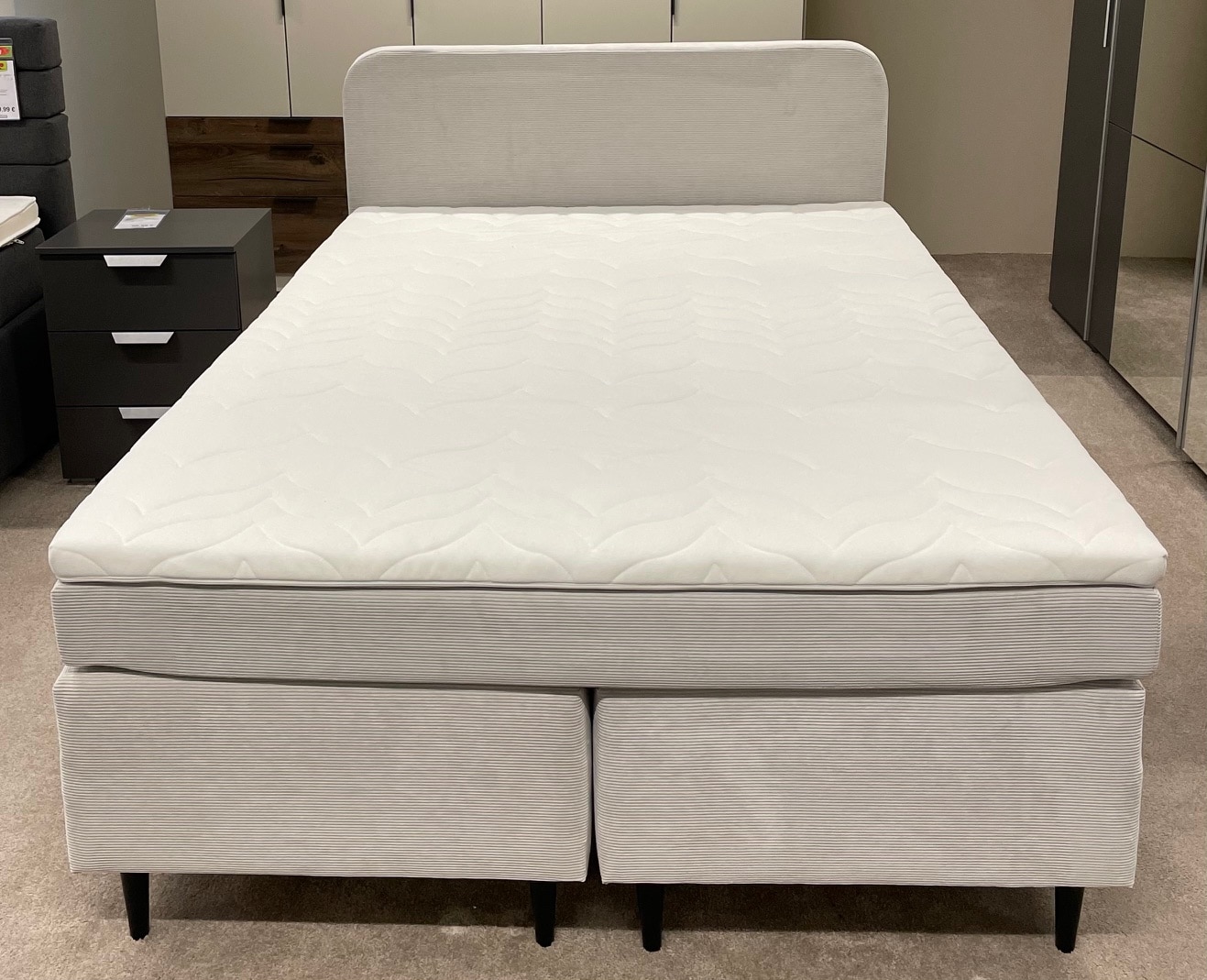Das Boxspringbett Wilmine ist ein modernes Bett mit einem hellgrauen gepolsterten Rahmen und Kopfteil in einem Ausstellungsraum.