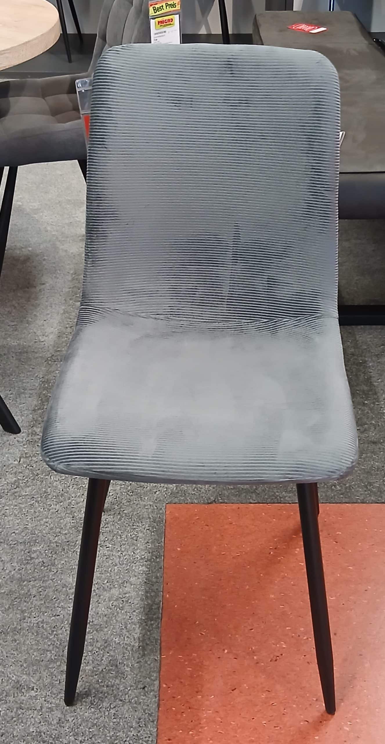 Vierfußstuhl Mike mit grauem Stoff und schwarzen Beinen auf einem grau-orangenen Teppich in einem Ausstellungsraum.
